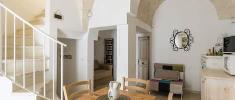 A dining area with a wooden table, chairs, and a vase. There is a staircase with white railings and a view of a living room with a sofa and a television.