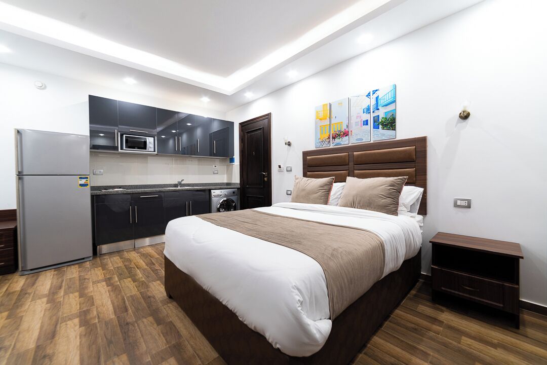 A modern bedroom with a large bed, a built-in kitchen with a refrigerator, microwave, and oven, and a wooden floor.