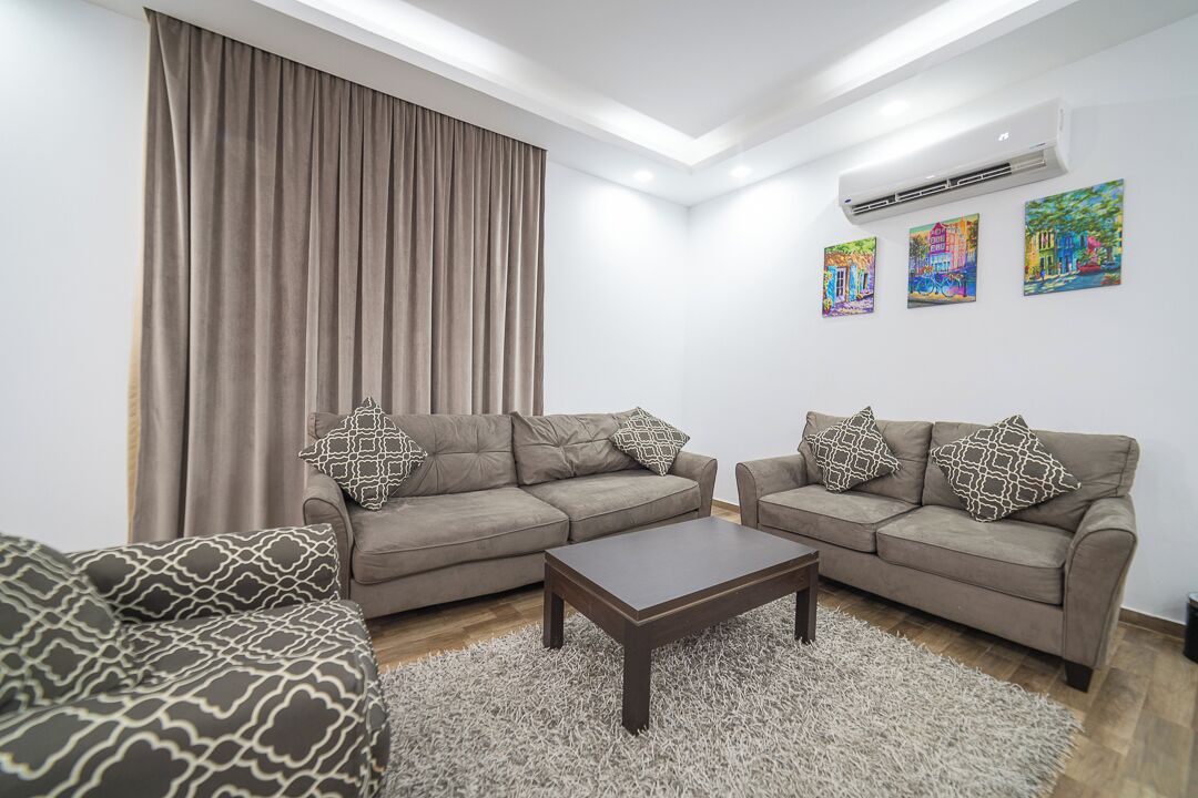A living room with two sofas, a coffee table, and a large window with curtains. There are three paintings on the wall.