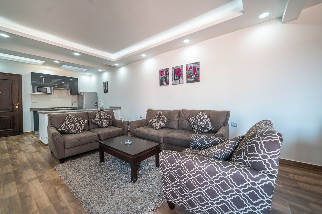 A modern living room with a sofa set, a coffee table, and a rug. There is a kitchen area visible in the background.