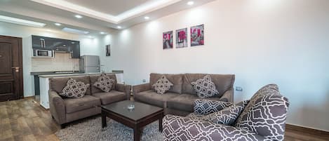 A modern living room with a sofa set, a coffee table, and a rug. There is a kitchen area visible in the background.