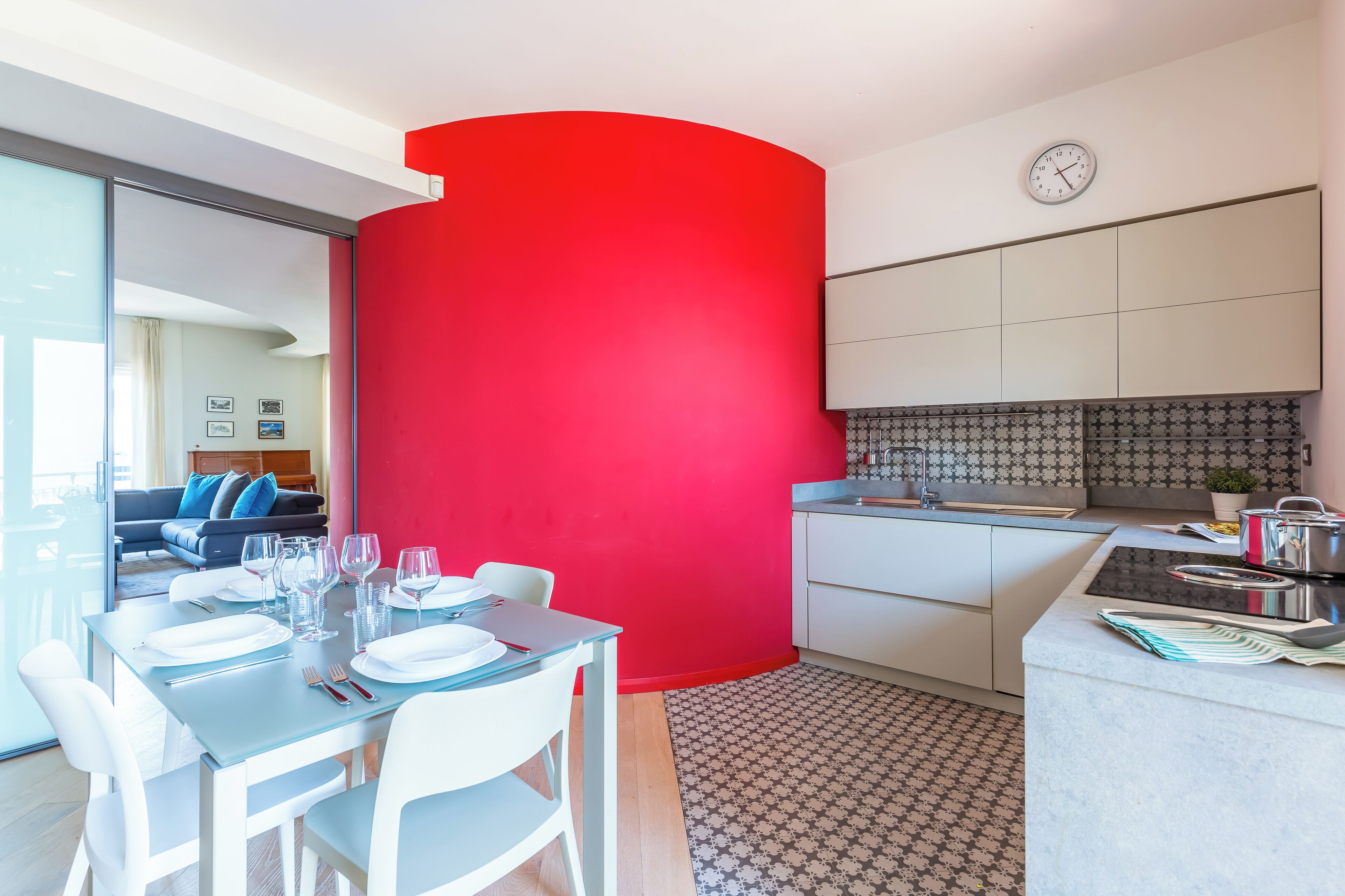 A modern kitchen with white cabinets, a red accent wall, and a dining area with white chairs and a glass table.