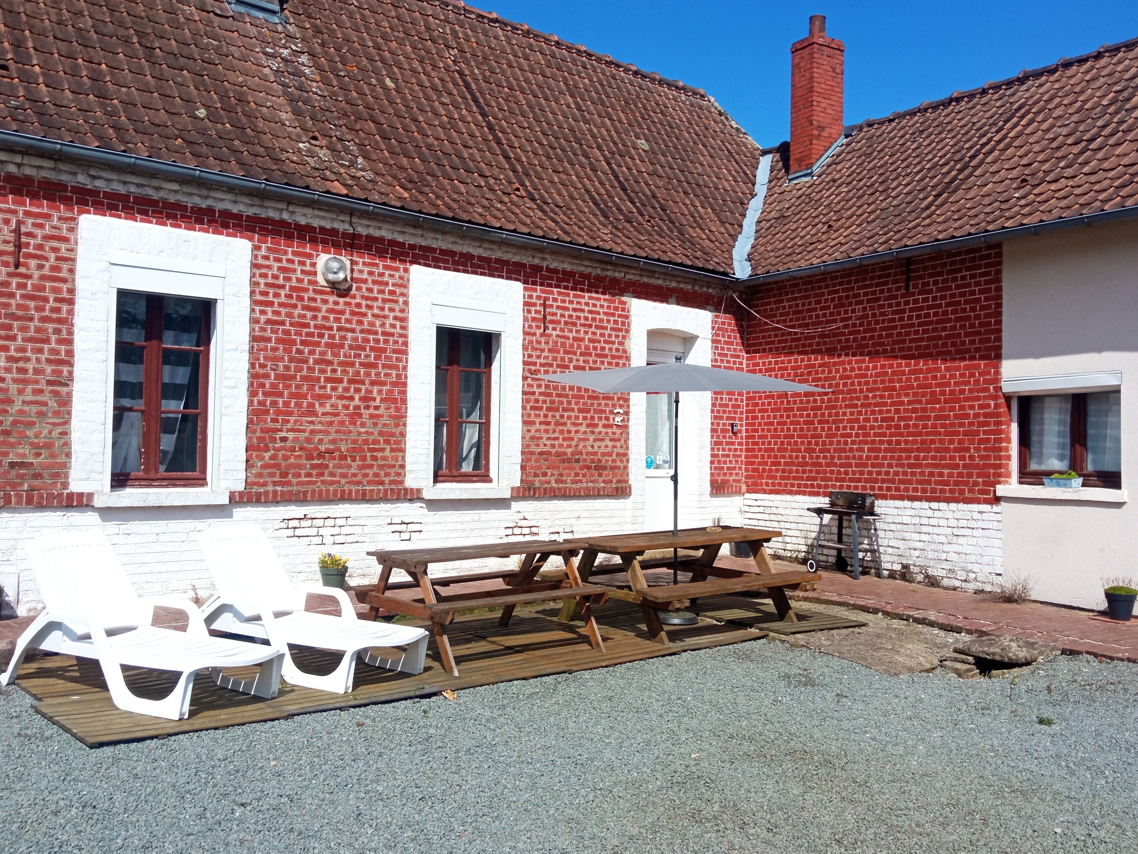 Een rood bakstenen gebouw met witte details, een houten tafel met banken en een witte parasol.
