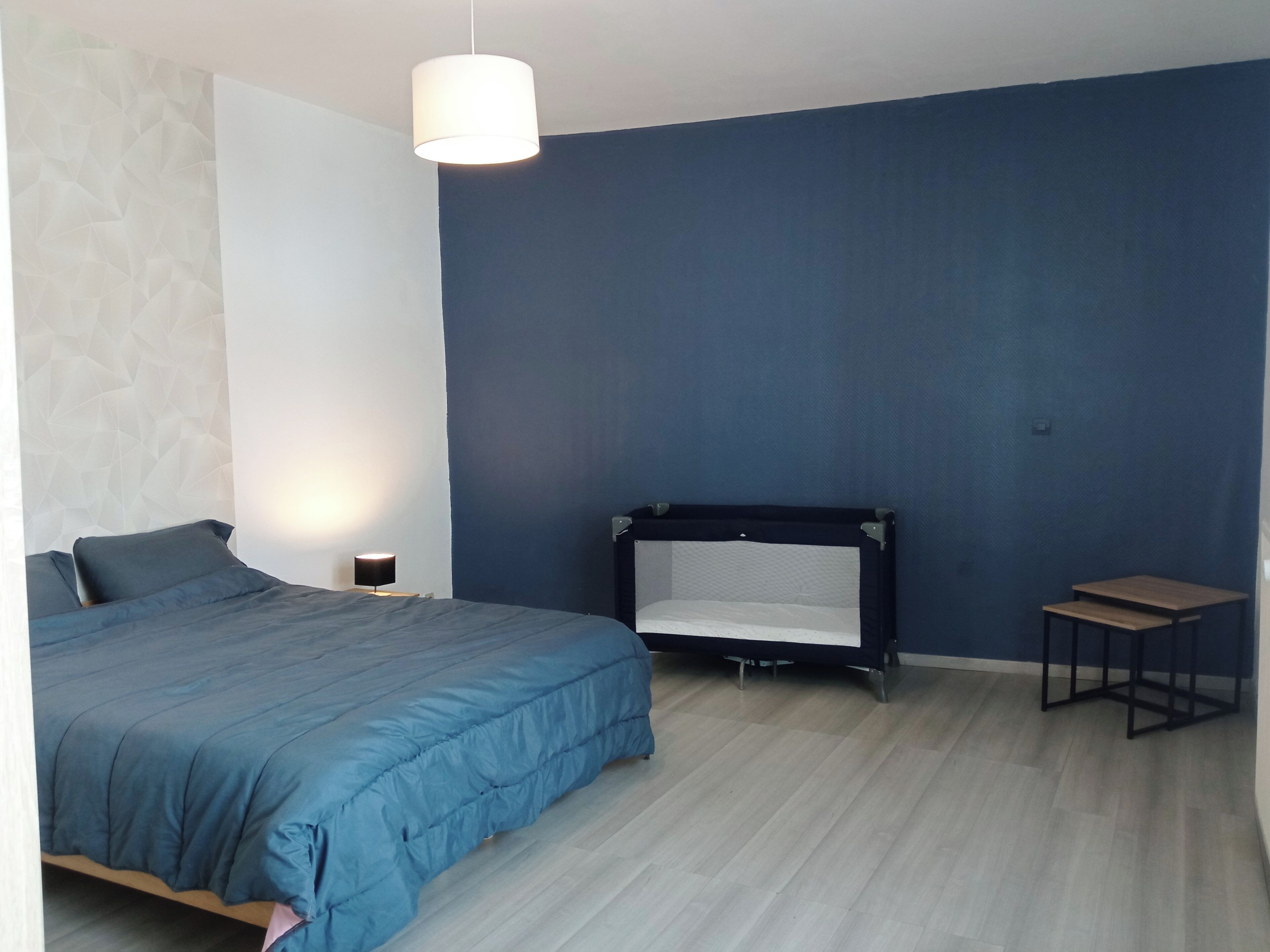 A bedroom with a bed, a bedside table, a hanging light, a bench, and a wall with a geometric pattern.