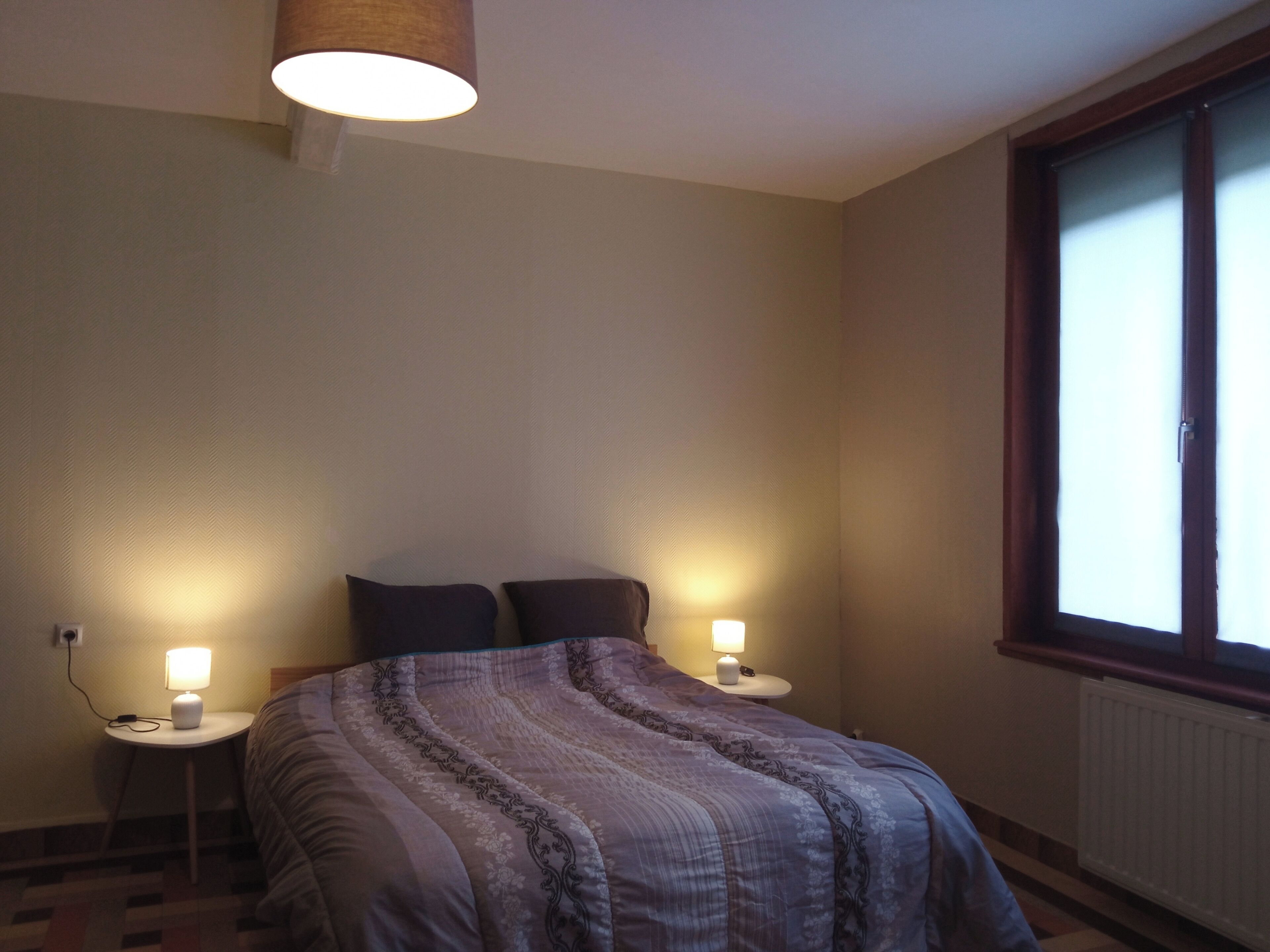 Ett sovrum med en säng, två sänglampor, ett fönster och ett element.