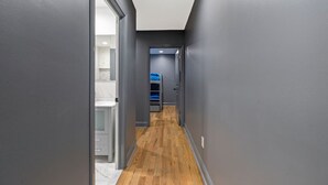 Un couloir étroit aux murs gris et au plancher en bois, qui mène à une pièce contenant un lit et une couverture bleue.