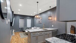 Une cuisine moderne avec des meubles gris, un plan de travail en marbre, une cuisinière encastrée, des luminaires suspendus et un petit coin salon avec un canapé gris et une fenêtre équipée de stores.