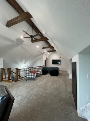Ein geräumiges Dachzimmer mit hoher Decke, freiliegenden Holzbalken, einem Deckenventilator und einem an der Wand befestigten Flachbildfernseher.