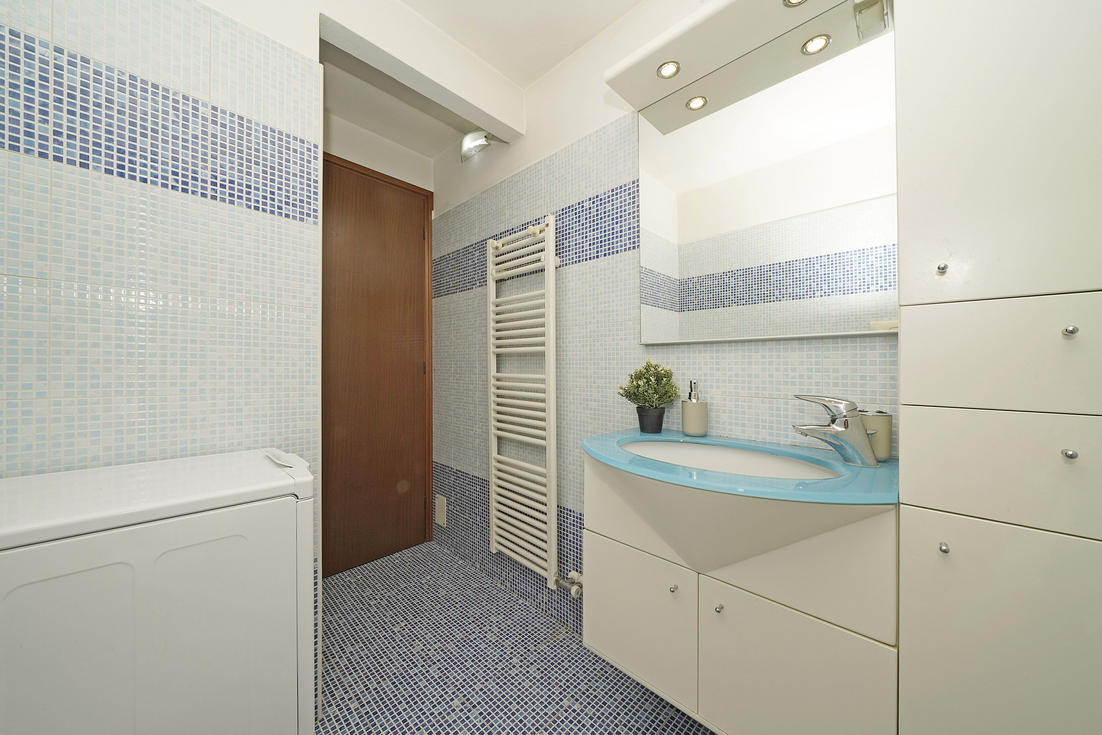 A bathroom with a white radiator, a blue-tiled sink, and a mirror above it.