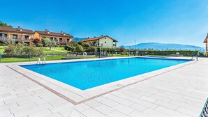 Ein rechteckiger Swimmingpool mit einer gefliesten Terrasse, umgeben von einem Zaun, mit Blick auf Wohnhäuser und Berge im Hintergrund.