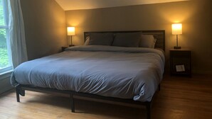 A bedroom with a bed, two bedside tables with lamps, a window with curtains, and a wooden floor.