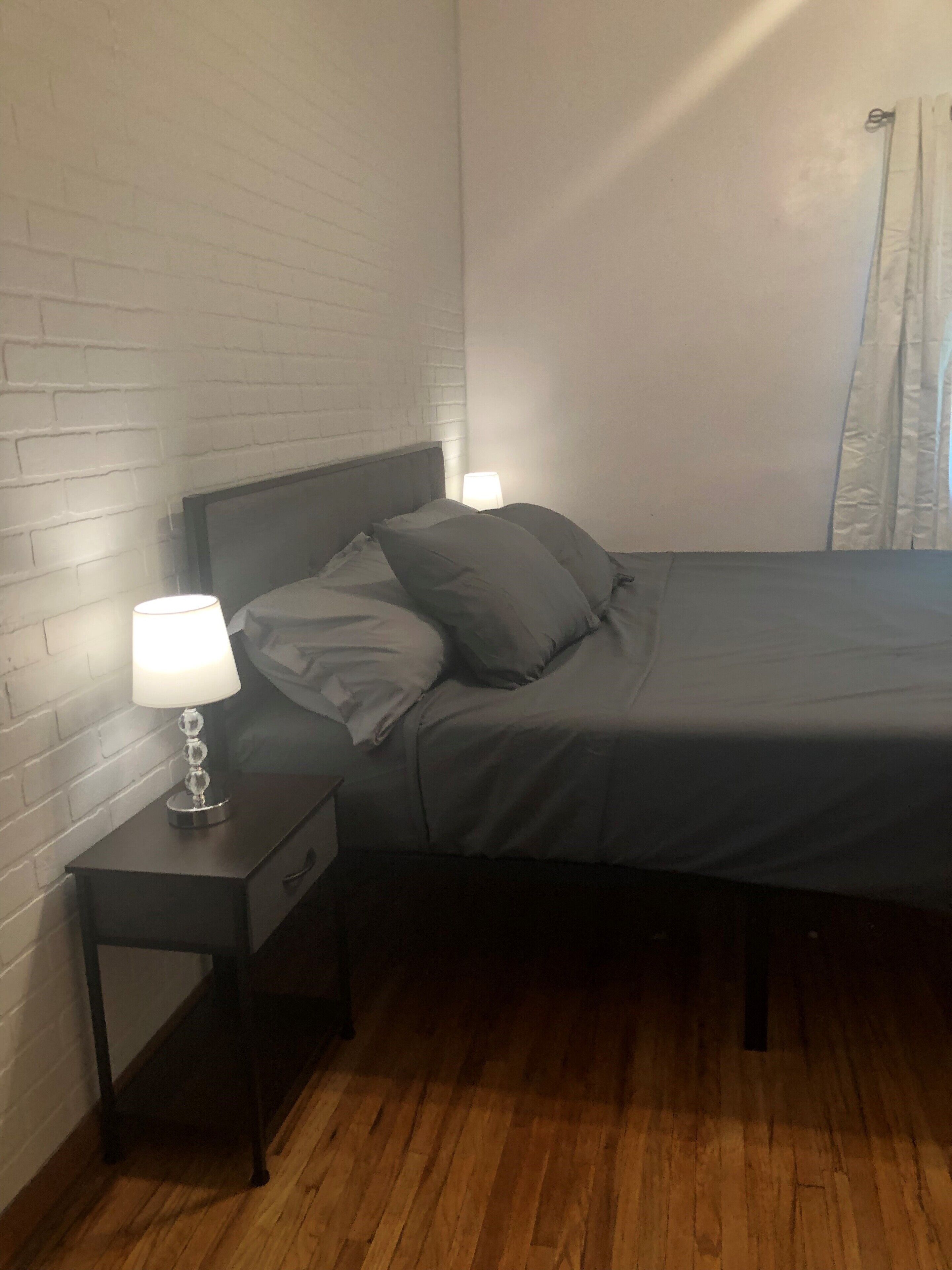 A bedroom with a wooden floor, a bed with grey bedding, a nightstand with a lamp, and a white brick wall.