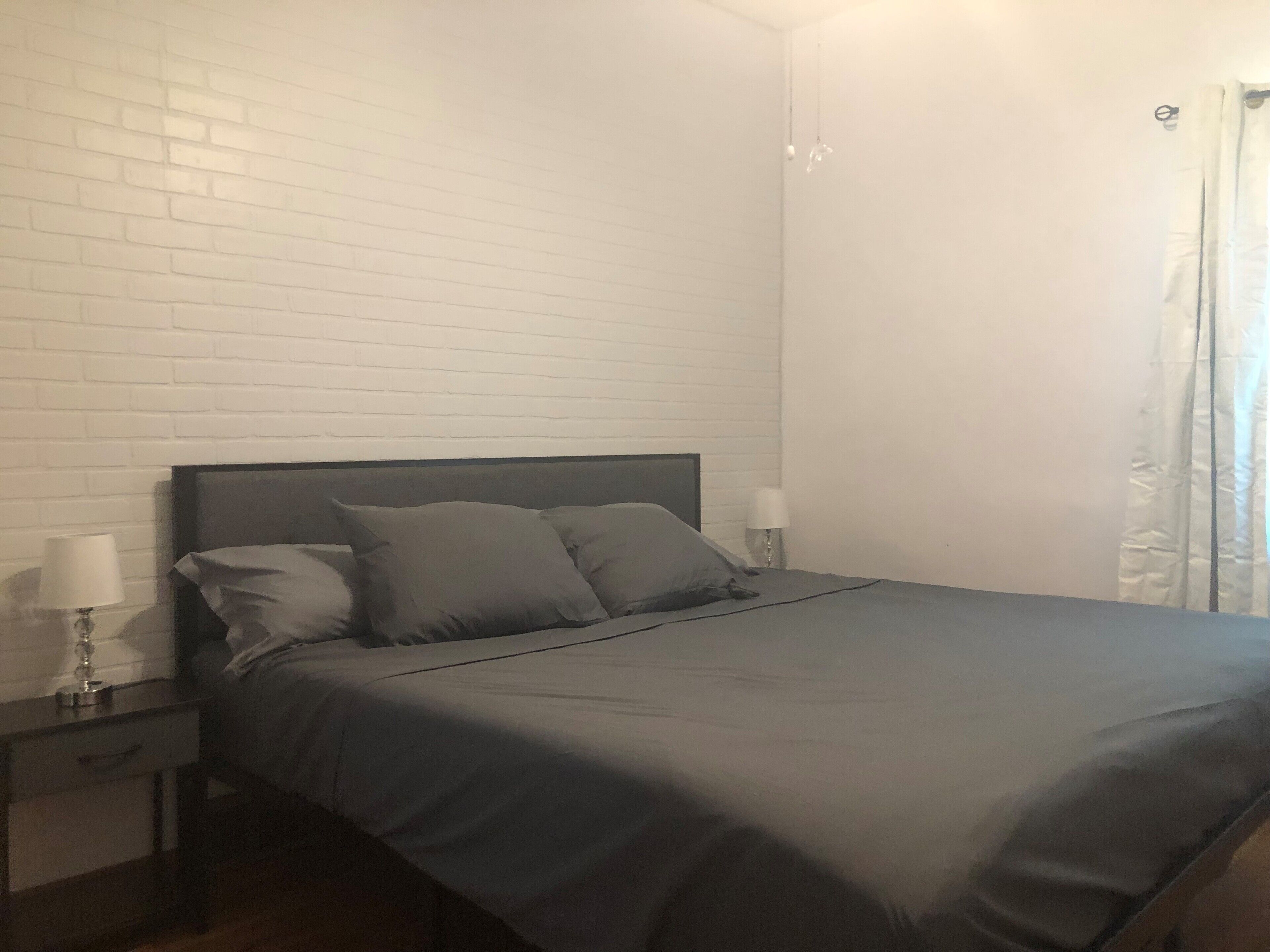 A neatly made bed with a grey bedspread and pillows, a dark headboard, and two bedside lamps on a nightstand.