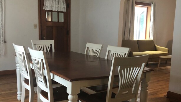 A dining room with a wooden table and white chairs, a ceiling fan, and a window with curtains.