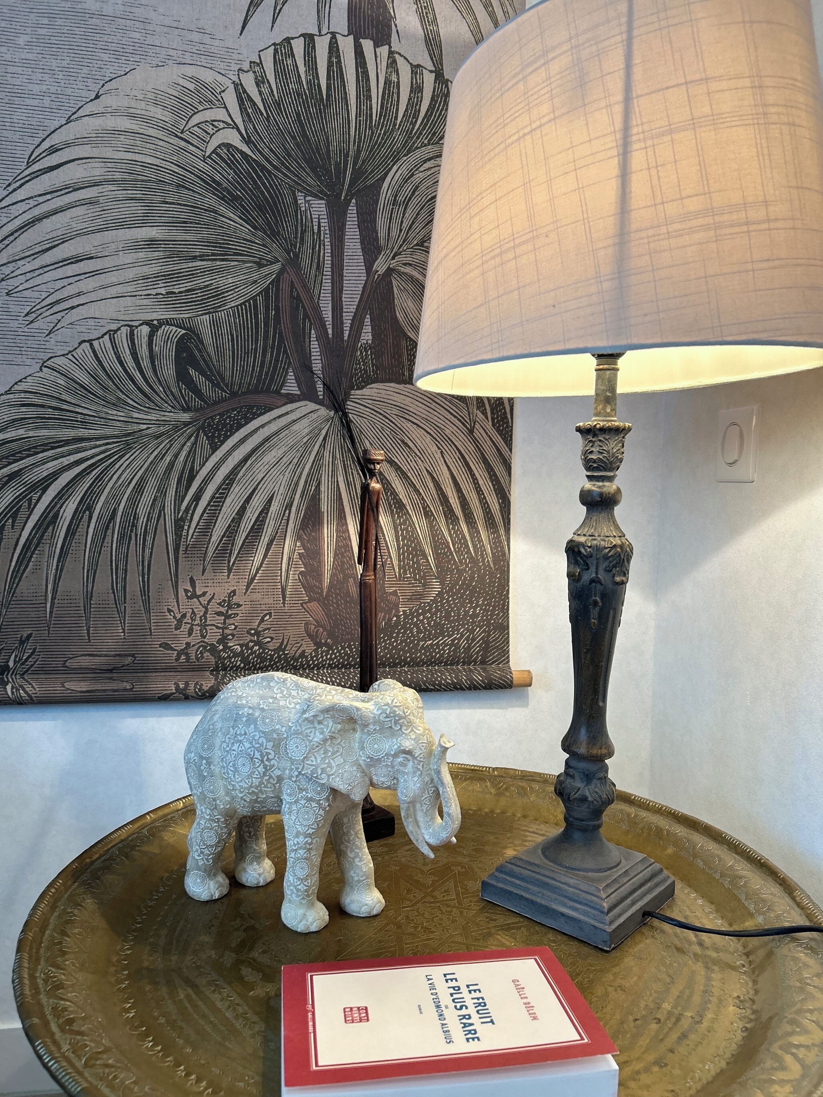 A lamp with a textured shade, a decorative elephant figurine, and a book titled "Le Fruit Le Plus Rare" on a tray.