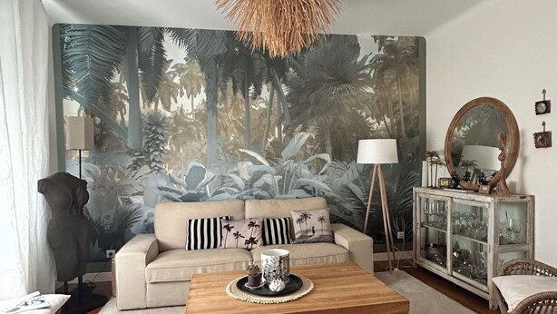 Un soggiorno con un'ampia murale tropicale, un divano con cuscini a righe, un tavolino in legno, poltrone in vimini, un mobile vetrina e un lampadario.