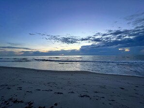 Ein Strand mit Fußspuren, die zum Wasser führen, bewölkter Himmel und eine untergehende oder aufgehende Sonne.