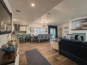 A spacious living area with a dark blue sofa, a dining table with chairs, a kitchen in the background, and a view of the outdoors through a window.