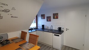 Una cuina moderna amb armaris blancs, tauleig negre i un rentaplats. Hi ha dues cadires de vimetina i una taula de dinar petita. La cuina té una finestra i obra d'art que representa utensilis de cuina.