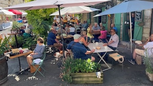 Ein Straßencafé mit Tischen und Sonnenschirmen, umgeben von Grünflächen und Steinbauten.