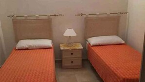 Una habitación pequeña con dos camas individuales, cada una con colcha naranja y almohadas blancas. Hay una mesita de noche con una lámpara entre las camas.