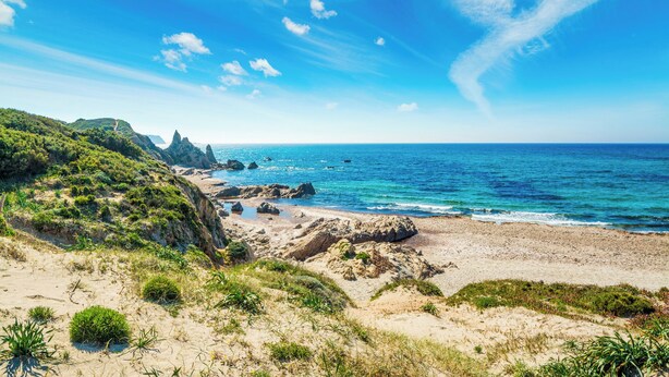 A coastal landscape with a sandy beach, rocky outcrops, and lush greenery.