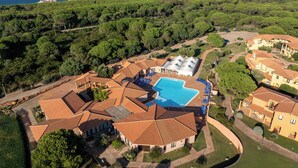 An aerial view of a resort with a central swimming pool, surrounded by villas with terracotta roofs, set against a backdrop of dense greenery and the sea.