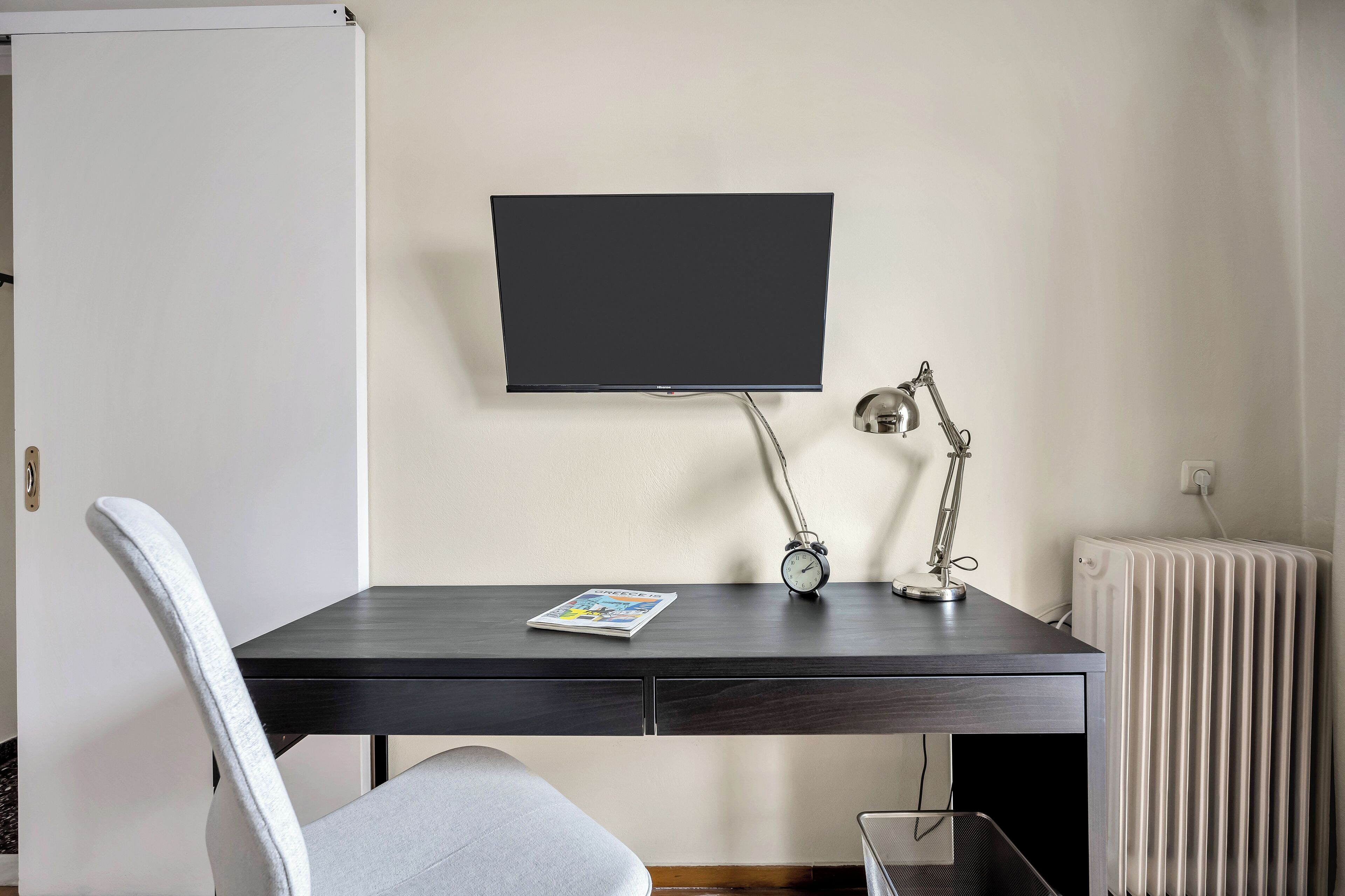 Une pièce moderne avec un bureau en bois foncé, une chaise blanche, un téléviseur fixé au mur, une lampe de bureau et un radiateur.
