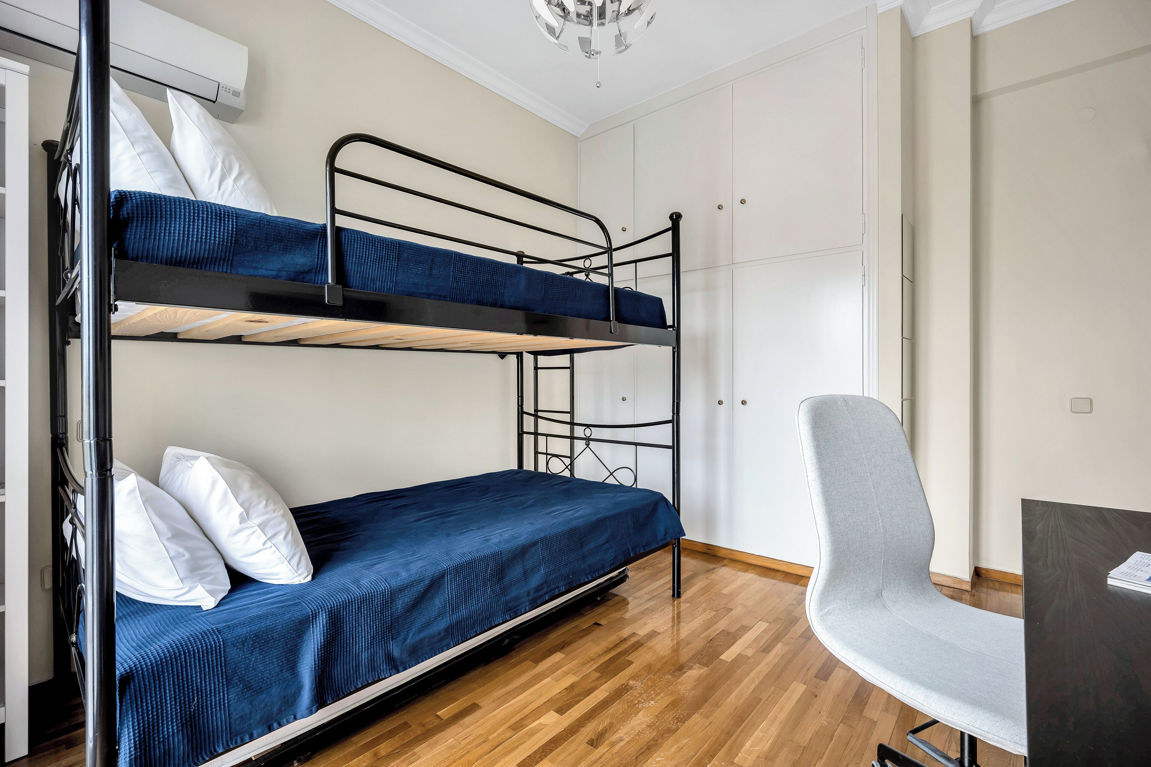 A bunk bed room with a desk and chair, white walls, and wooden flooring.