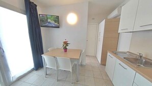 Et lille, moderne køkken med hvide skabe, en træbordplade og en vask. Der er et spisebord med to stole og en potteplante. Et fjernsyn er monteret på væggen over bordet.