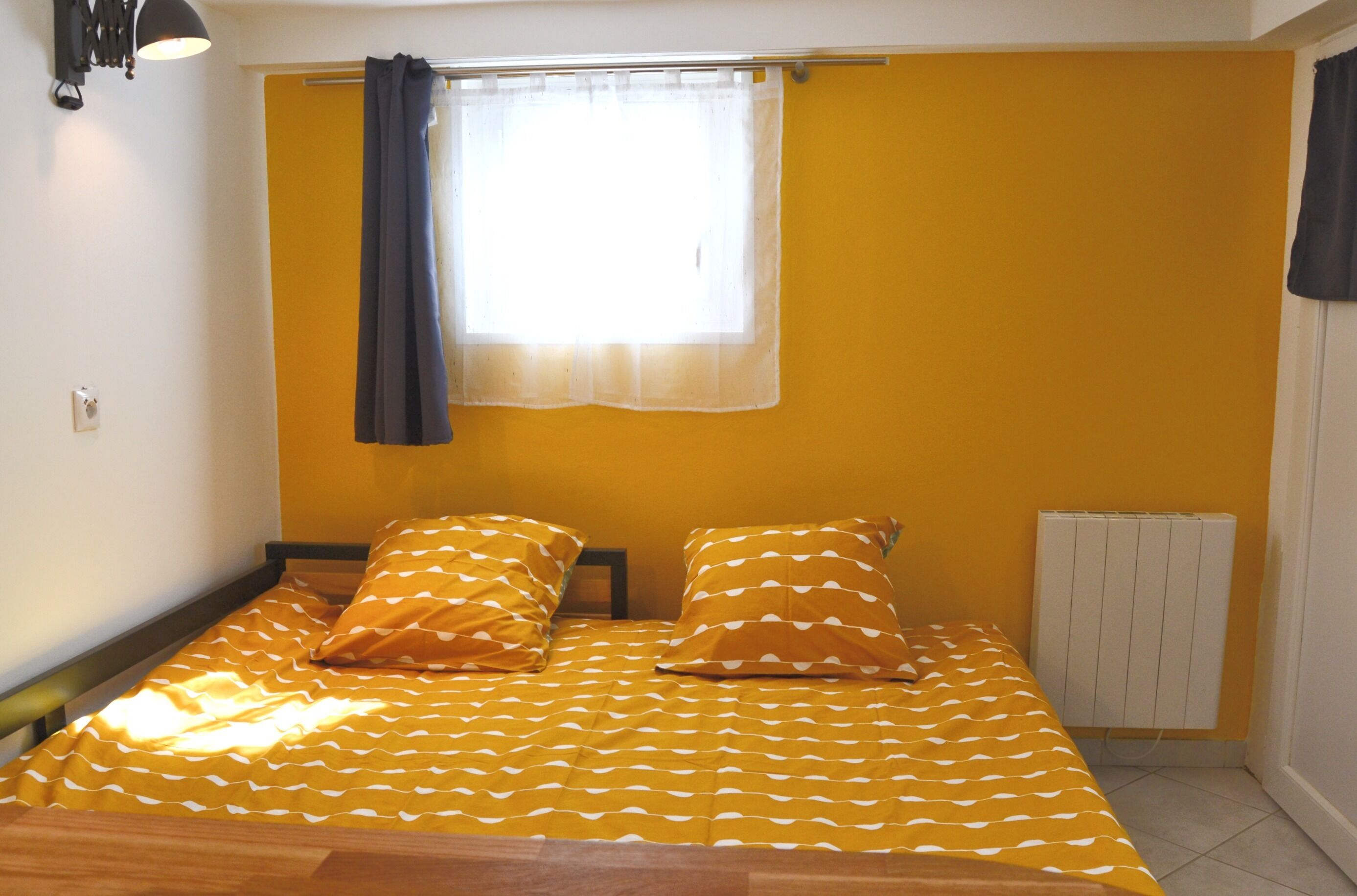 A room with a bed, a window with curtains, a radiator, and a wall-mounted light fixture.