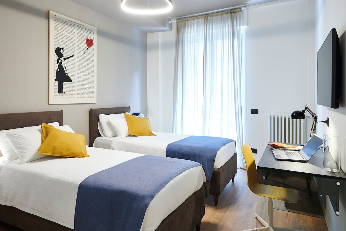 A hotel room with two beds, a desk with a laptop, a TV, and a window with curtains.