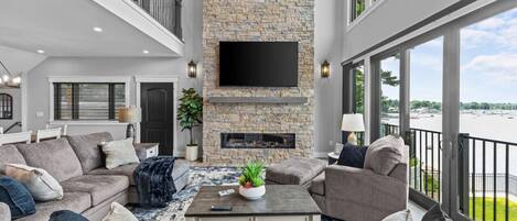 A spacious living room with a stone fireplace, large windows, and a balcony overlooking a body of water.