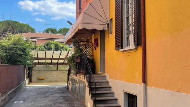 A yellow building with a staircase leading to a door, a covered outdoor area with plants, and a clear sky. - Elle Home: comfort in Bologna (Bologna)