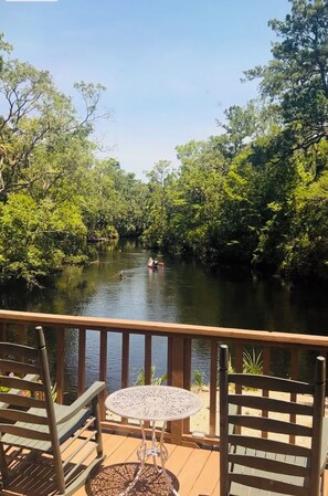 A wooden deck with a table and chairs overlooking a calm river surrounded by dense greenery. - The River House - Island Getaway on the Steinhatchee River (Steinhatchee)