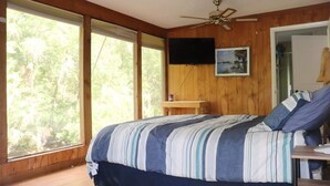 A bedroom with a large bed, wooden walls, a ceiling fan, a TV mounted on the wall, and a window with a view of trees. - The River House - Island Getaway on the Steinhatchee River (Steinhatchee)