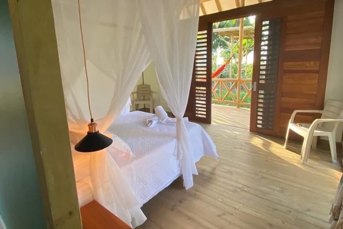 Quarto com cama sob dossel, piso de madeira e vista para um jardim por entre venezianas abertas.