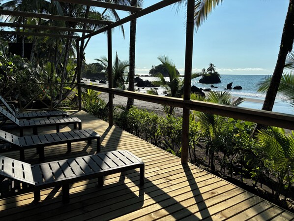 A wooden deck with lounge chairs overlooking a beach with palm trees and rocky outcrops.