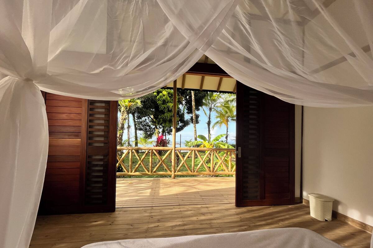 Quarto com cama, piso de madeira e vista para um deck de madeira e vegetação tropical.