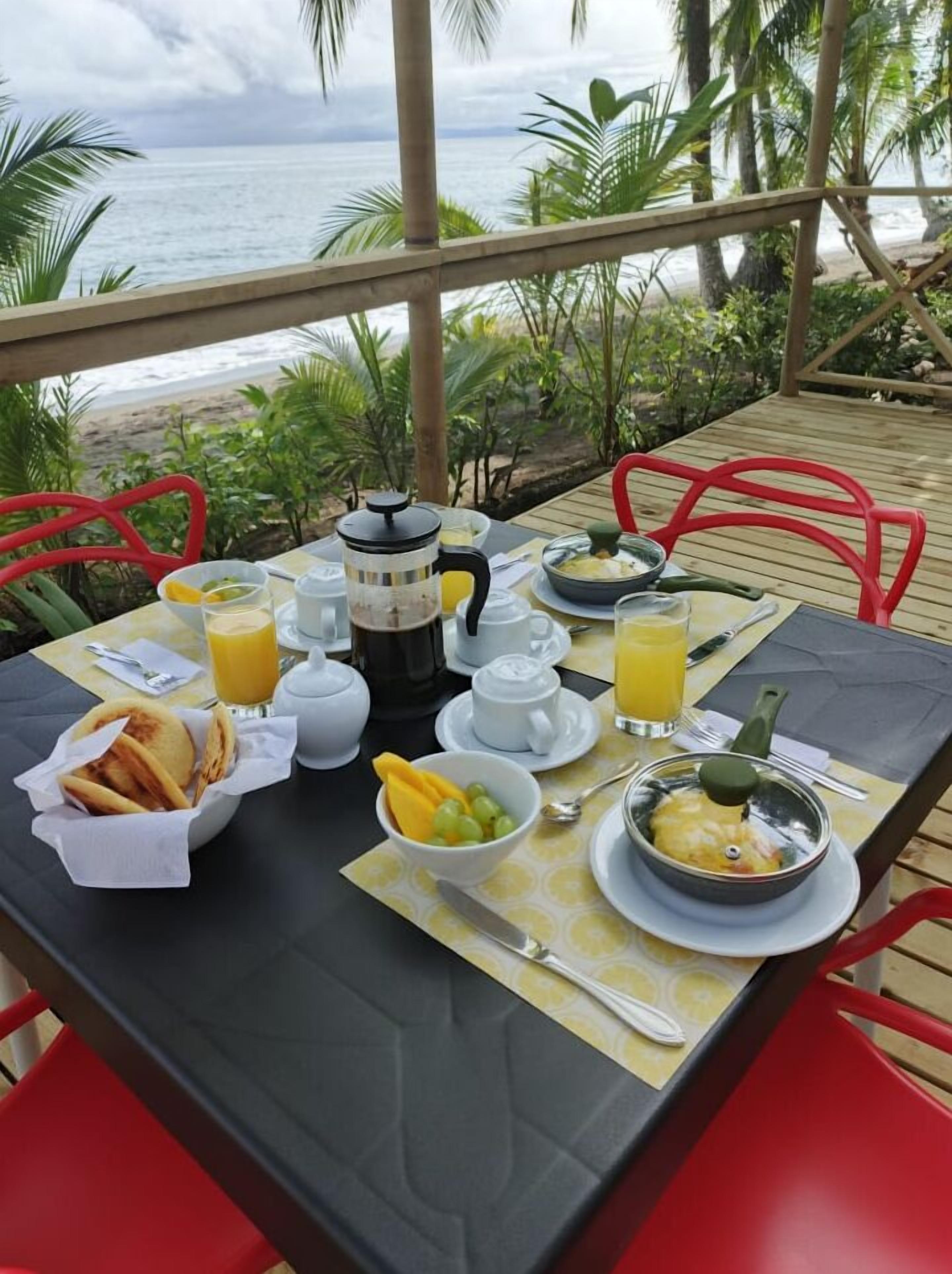 A breakfast table set on a wooden deck overlooking the ocean, with a view of palm trees and the sea.