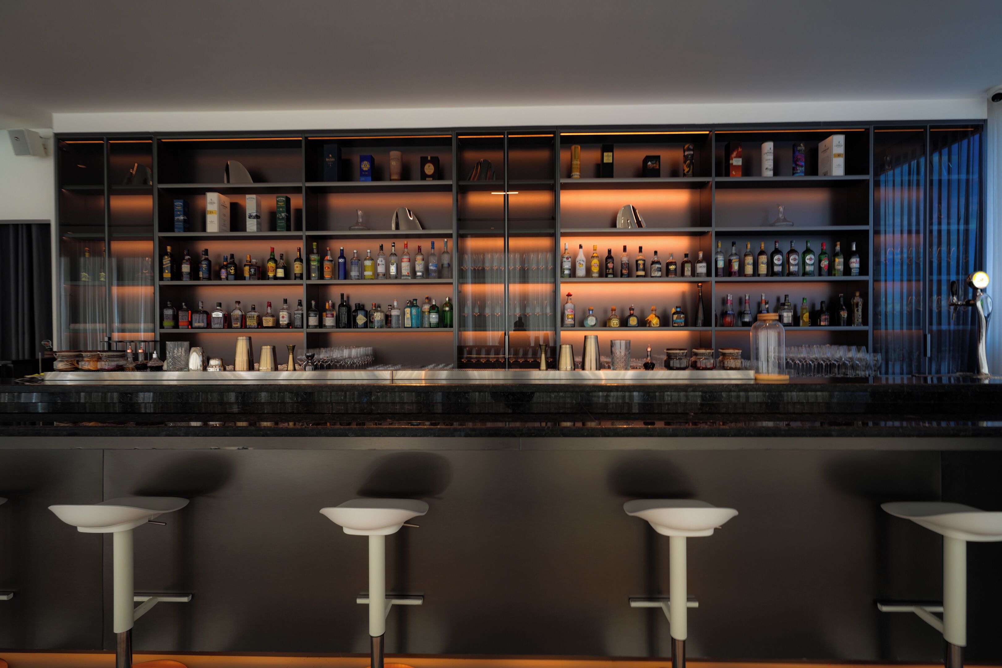 A bar area with shelves of liquor bottles and glasses, and three white bar stools.
