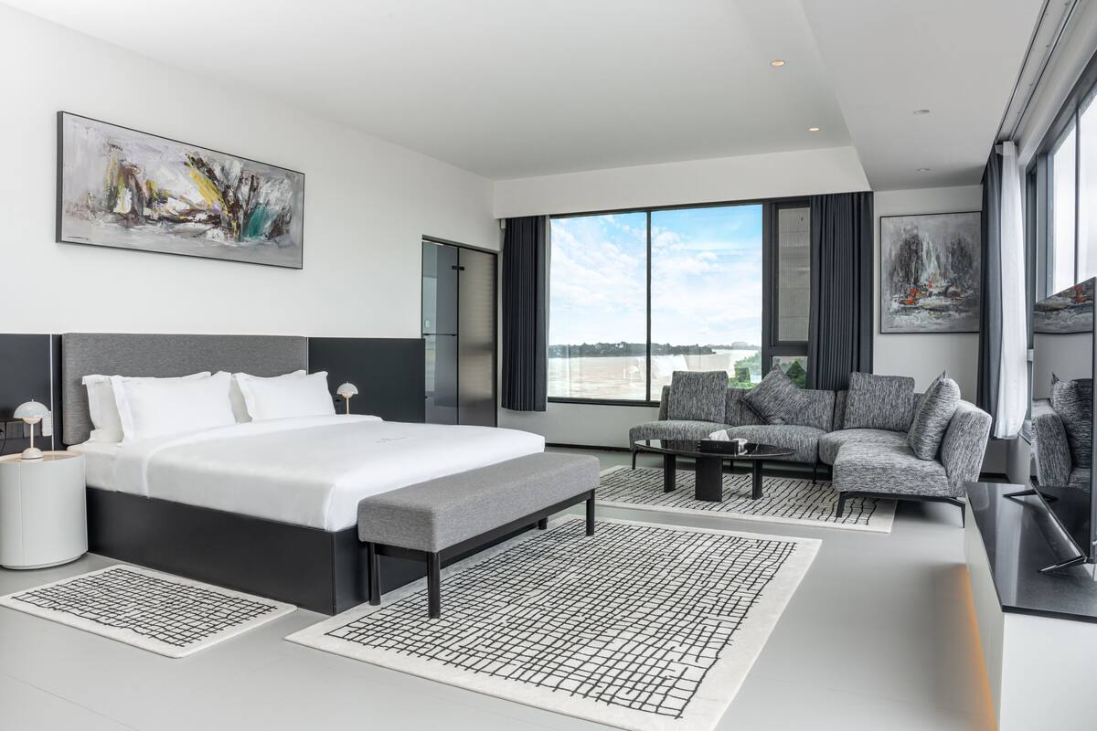A modern hotel room with a large bed, grey sofa, and a view of the city.