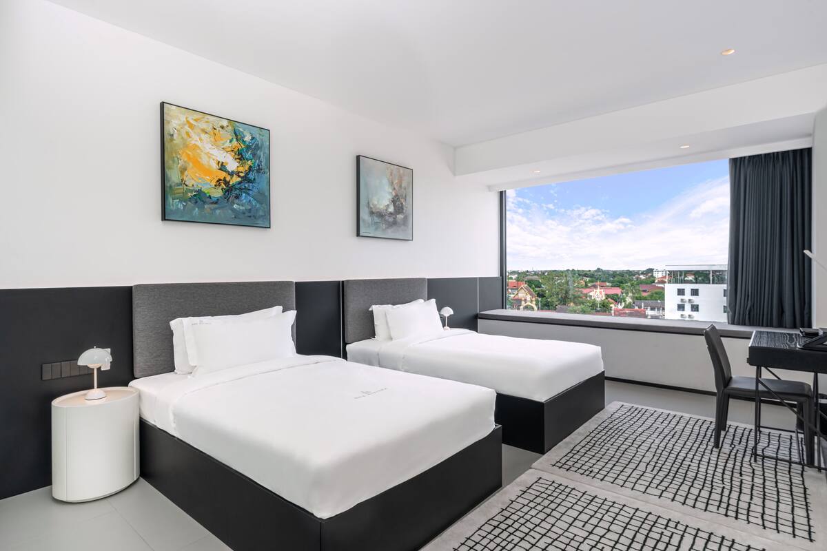 A modern hotel room with two beds, a desk, and a large window offering a city view.