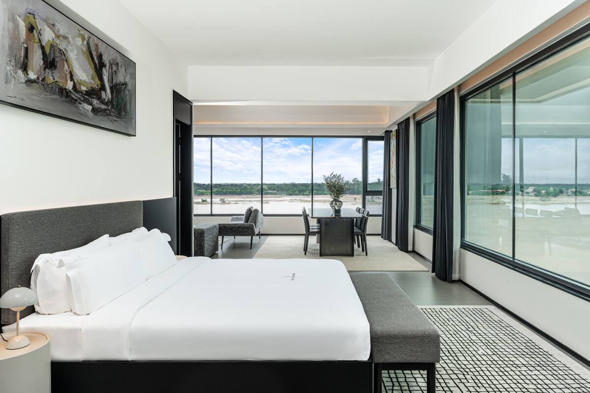 A modern hotel room with a large bed, a dining area with a table and chairs, and a balcony with a view of the landscape.