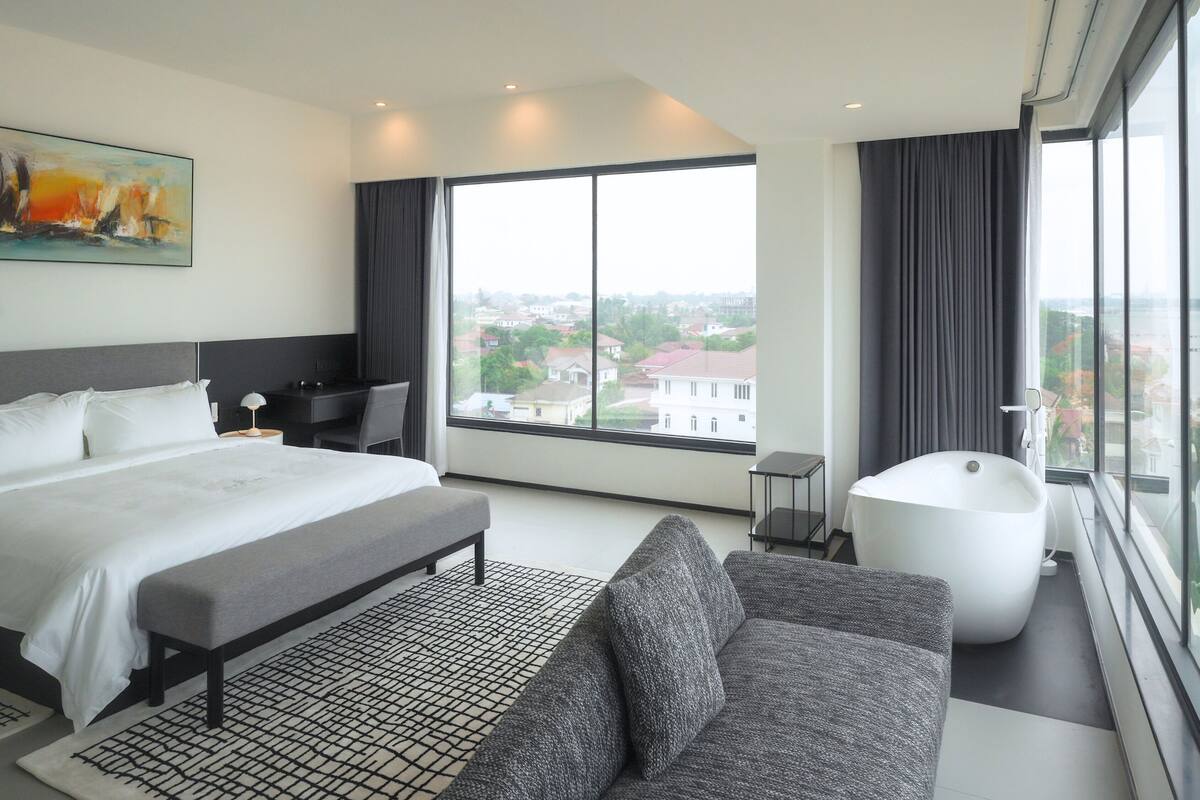A modern hotel room with a large bed, a sofa, a bathtub, and a city view.