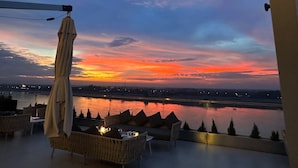 Une terrasse avec des sièges extérieurs et une vue sur un coucher de soleil se reflétant sur une étendue d’eau.