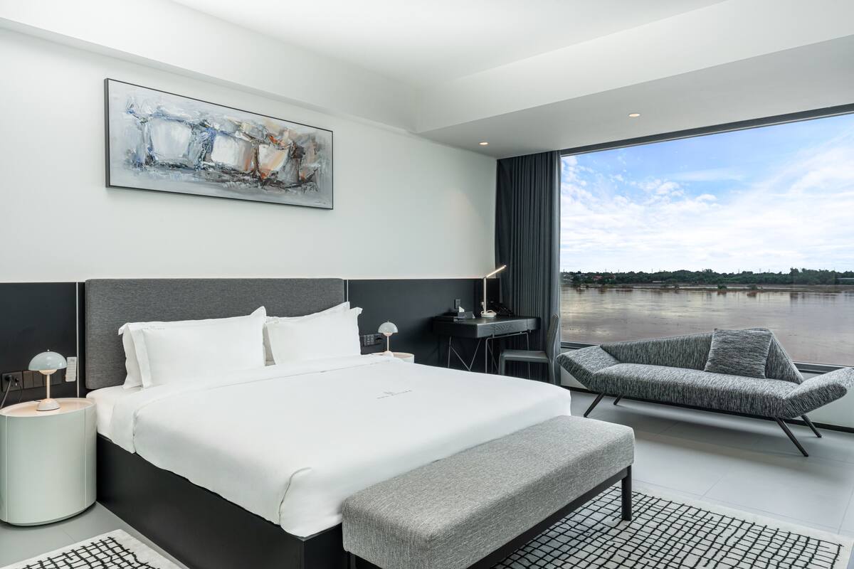 A modern hotel room with a large bed, a gray sofa, a desk, and a view of a body of water.