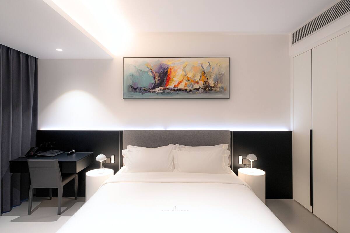 A modern hotel room with a large bed, a desk with a chair, and a painting on the wall.