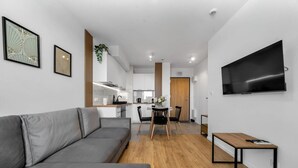 A modern living room with a grey sofa, a wooden dining table with chairs, a TV mounted on the wall, and a kitchen in the background. - 7th Floor Warsaw Włochy Apt - Balcony, Workspace - by Rentujemy (Warszawa)
