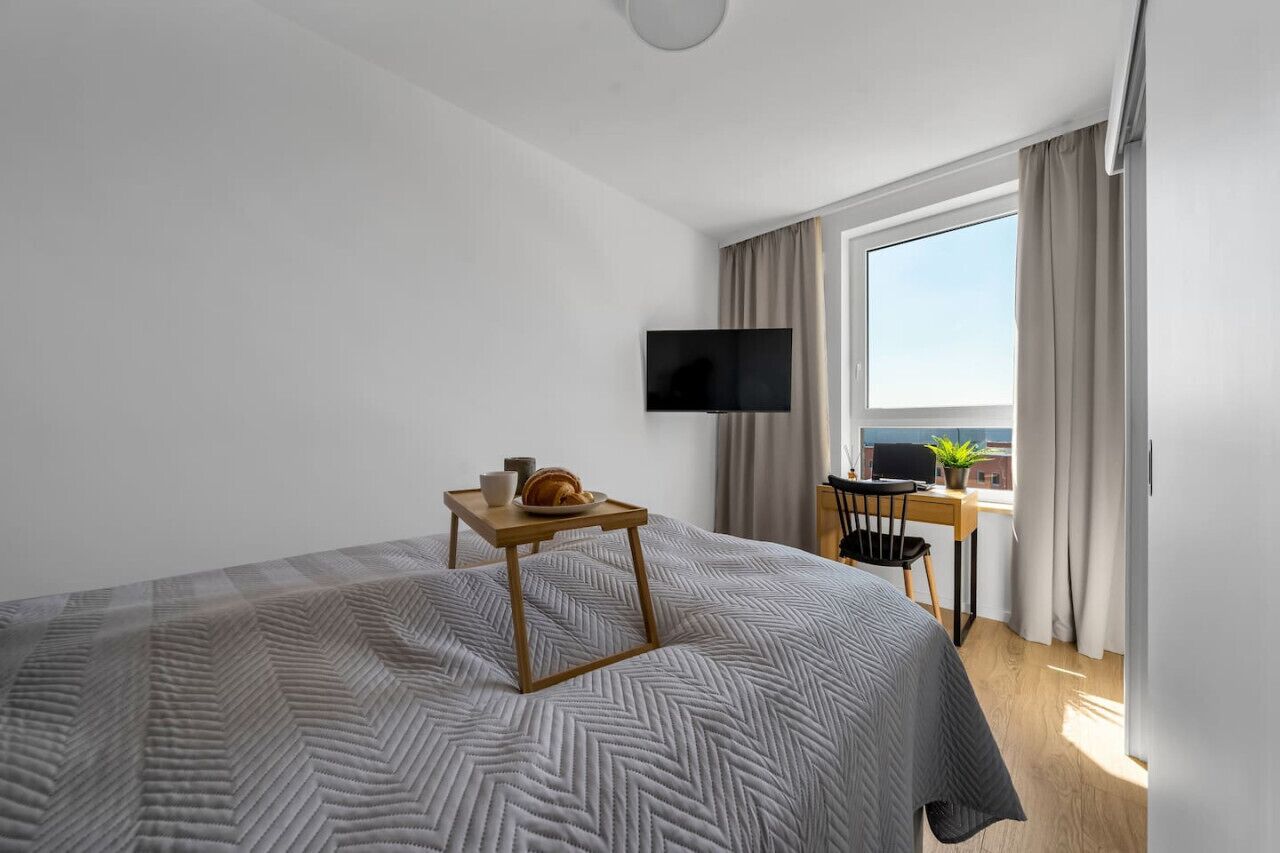 A hotel room with a large bed, a small table with a croissant, a TV on the wall, and a balcony with a view.